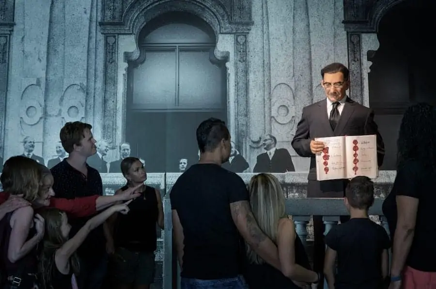 A wax figure of a man holding an open book stands on a stage in front of an image of a historical building. A group of people, including children and adults, stand below, observe the scene and point to it. 