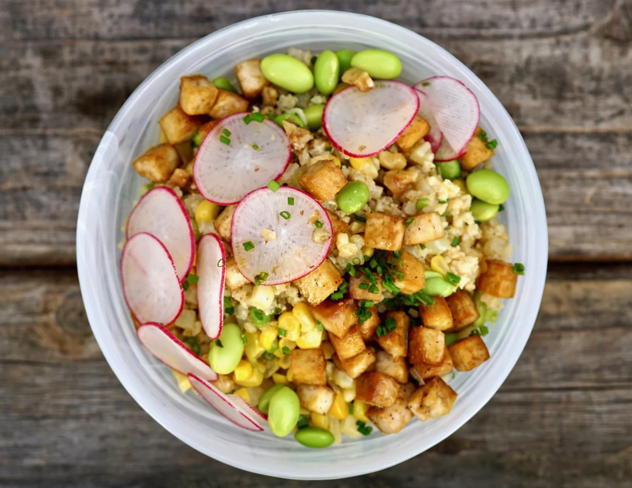 Eine Schüssel gebratener Reis, belegt mit Tofuwürfeln, Radieschenscheiben, Mais und Edamame. Das Gericht wird mit gehacktem Schnittlauch garniert und in einer weißen Schüssel auf einem rustikalen Holztisch serviert.