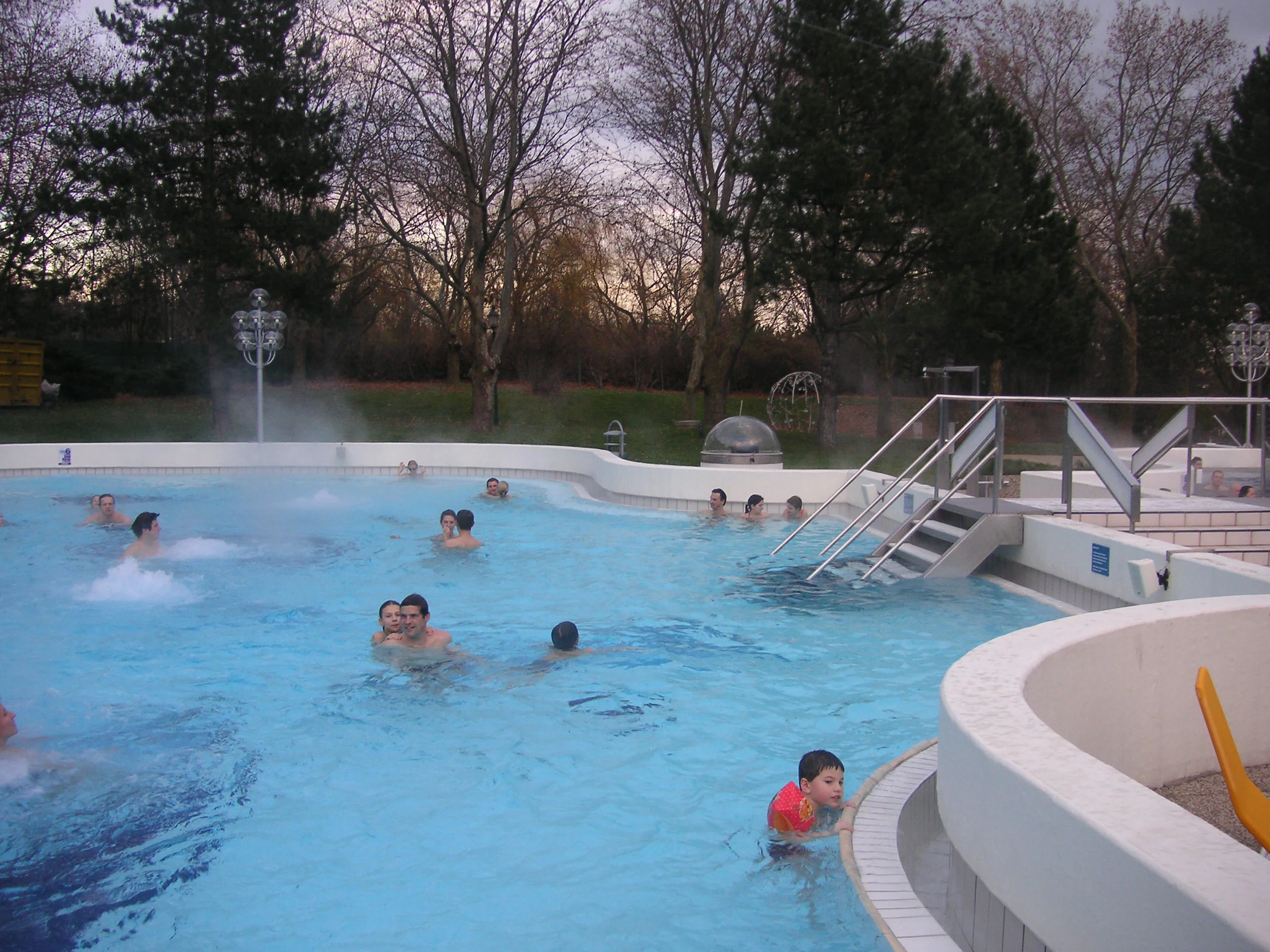 Menschen genießen ein Bad in einem von Bäumen umgebenen Außenpool. Einige spielen und schwimmen, während andere am Beckenrand entspannen. Der Himmel ist bewölkt und die Umgebung bietet eine ruhige, parkähnliche Umgebung.