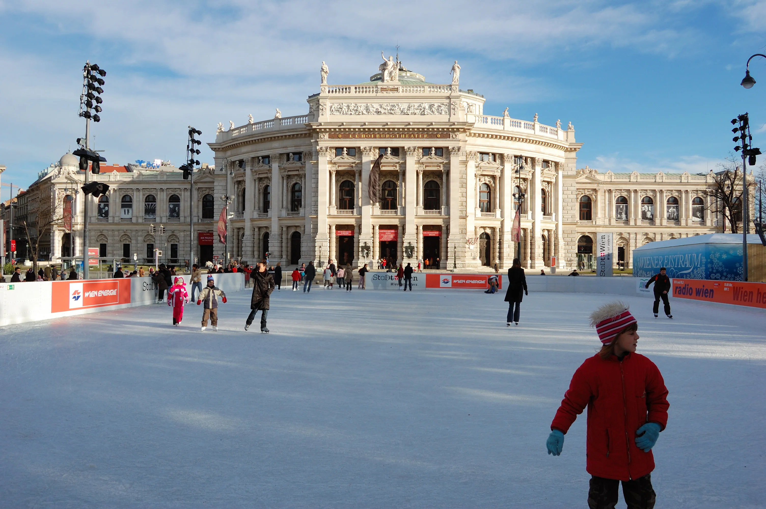 Menschen genießen das Schlittschuhlaufen auf einer großen Eisbahn vor einem kunstvollen historischen Gebäude mit Säulen und Statuen. Der Himmel ist klar und blau und einige Menschen tragen Winterkleidung wie Mäntel und Mützen.