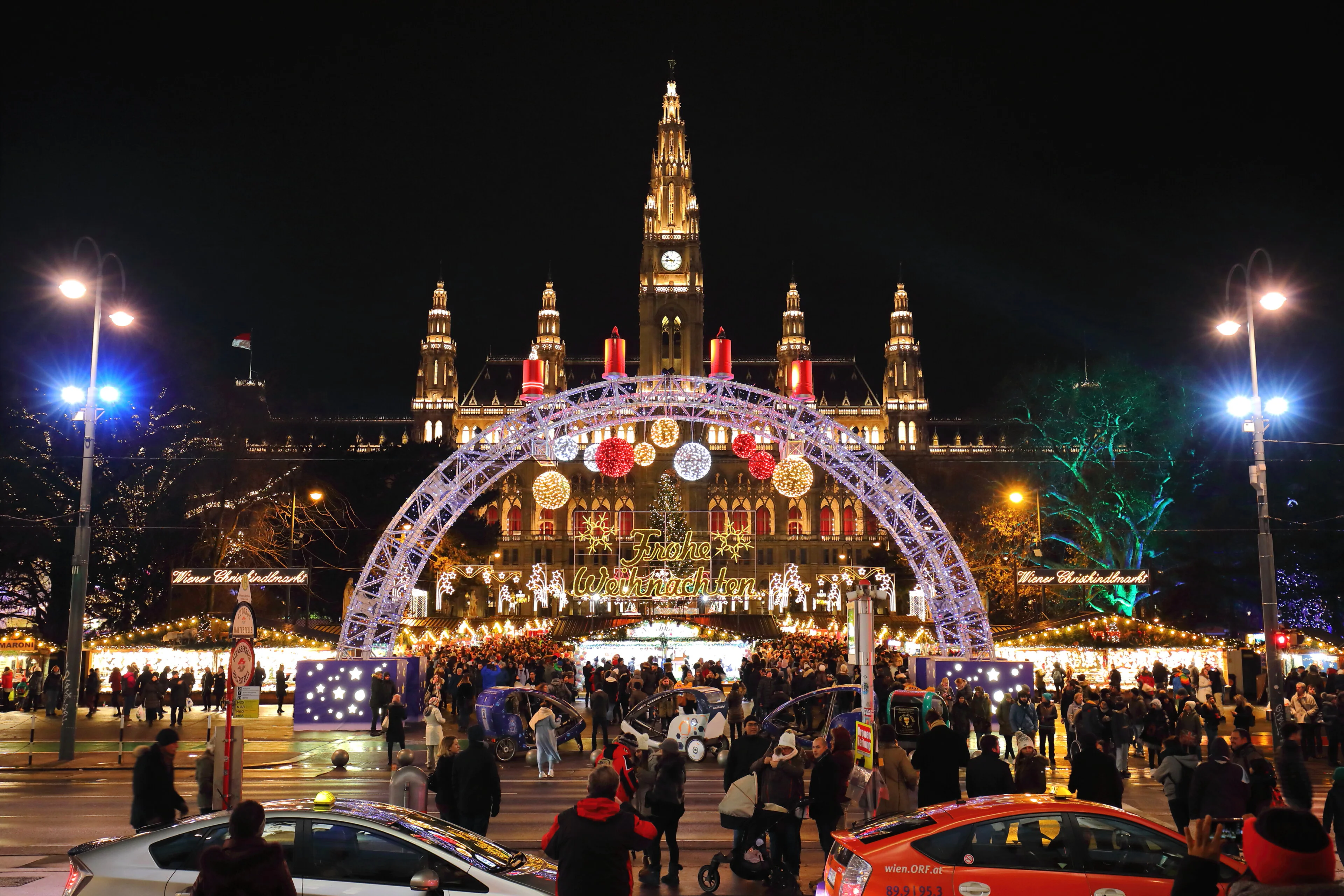 Ein geschäftiger Weihnachtsmarkt bei Nacht vor einem prächtigen, beleuchteten Gebäude. Ein großer Bogen mit festlichen Ornamenten und „Frohe Weihnachten“-Lichtern umrahmt die Szene, während zahlreiche Besucher und Straßenstände für eine lebendige Atmosphäre sorgen.