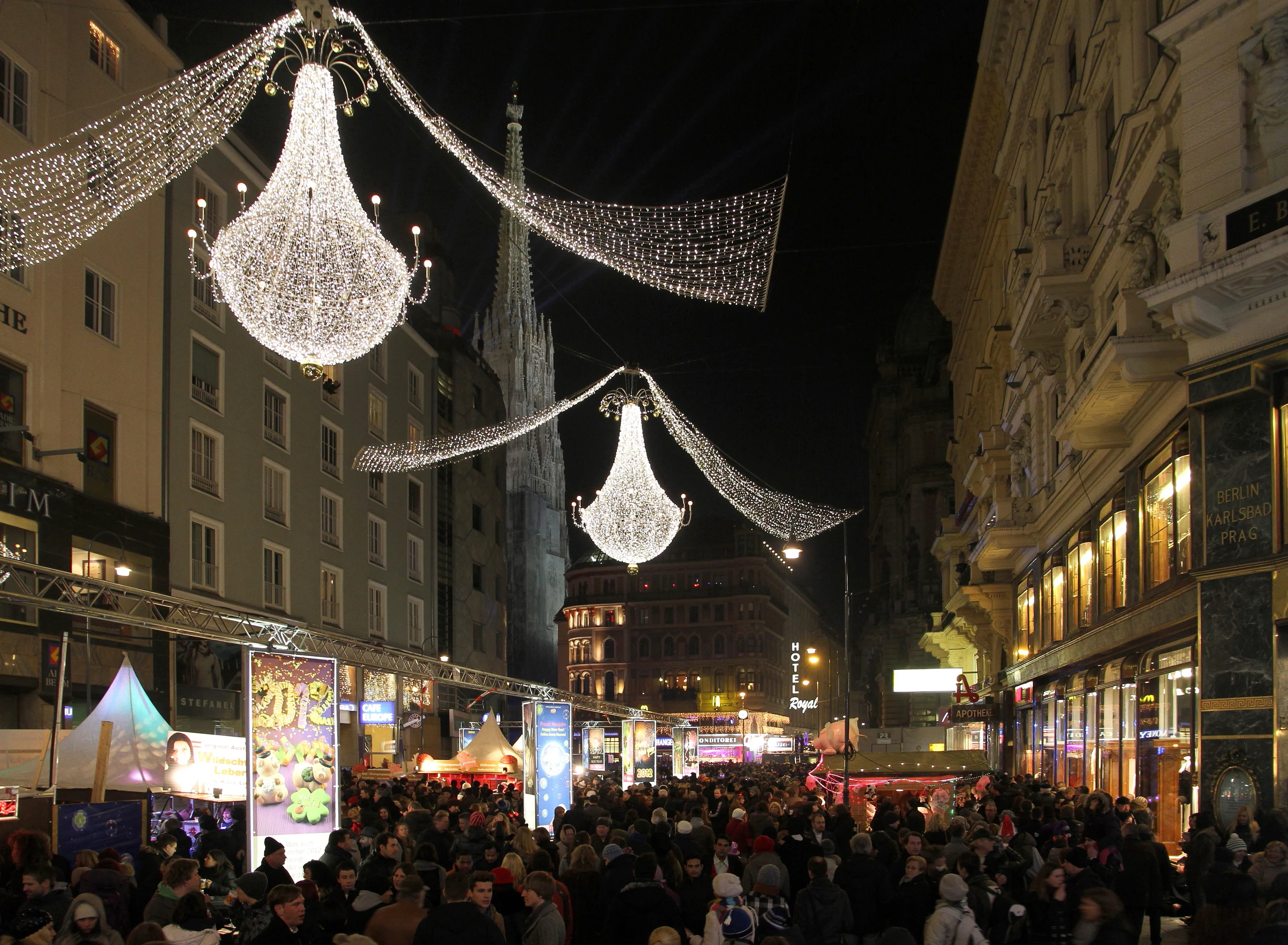 Eine belebte Stadtstraße ist nachts voller Menschen, die einen festlichen Markt besuchen. Große, beleuchtete Kronleuchter und Lichterketten hängen über der Menge. Gebäude sind dekoriert und Stände säumen die Straße und sorgen für eine lebendige Feiertagsatmosphäre.