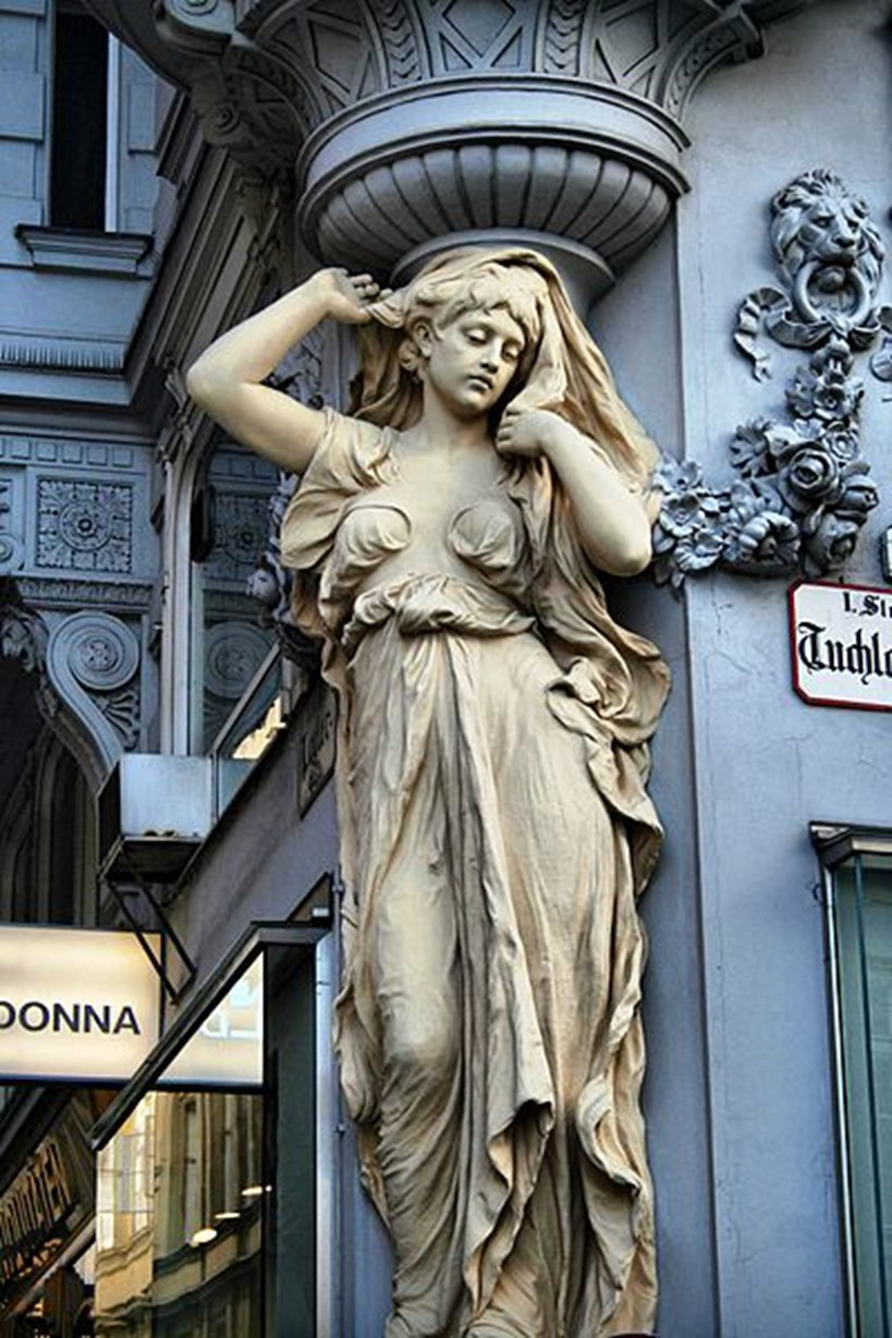 Steinstatue einer Frau in wallendem Gewand, die an einer kunstvoll verzierten architektonischen Säule lehnt. Die Statue hat ein kompliziertes Design mit detaillierten Schnitzereien oben und an der Seite und einem sichtbaren Ladenschild in der unteren linken Ecke.