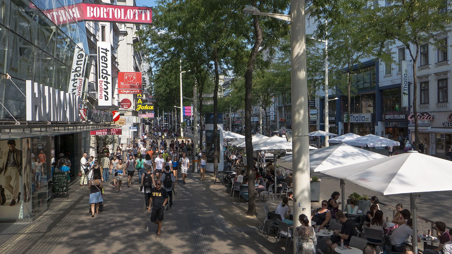 Eine geschäftige Straßenszene mit Menschen, die im Freien spazieren gehen und essen. Auf der linken Seite reihen sich Geschäfte aneinander, darunter H&M und Bortolotti. Auf der rechten Seite stehen weiße Sonnenschirme unter Tischen. Unter einem klaren Himmel sind Bäume und moderne Gebäude zu sehen.