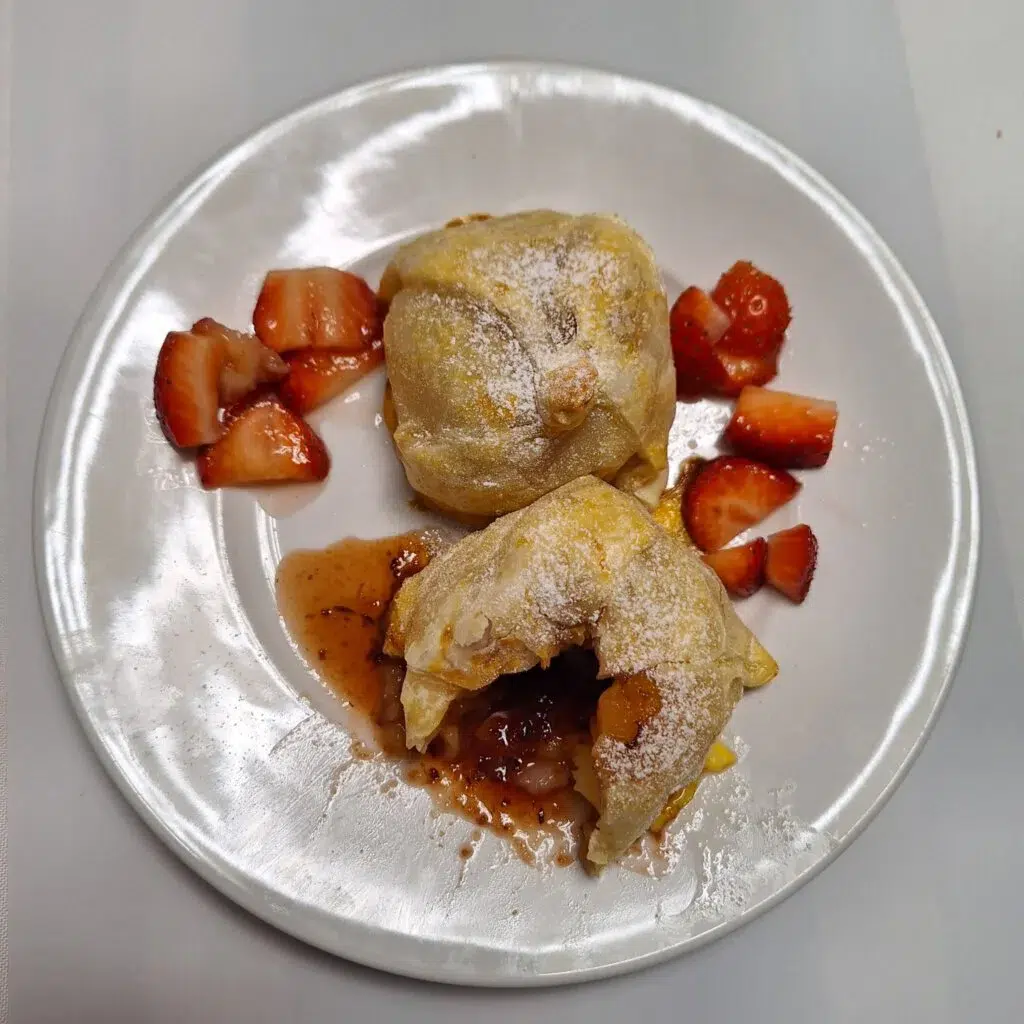 Ein weißer Teller mit zwei mit Puderzucker bestäubten Blätterteigtaschen, umgeben von geschnittenen Erdbeeren. Aus einem der Taschen ist etwas Füllung herausgelaufen, dazu ein Spritzer Erdbeersoße.