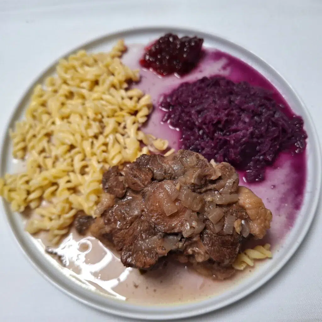 Ein Teller mit Rotini-Nudeln, Rotkohl, einer Portion gekochtem Fleisch mit Soße und einem Klecks Marmelade. Das Essen ist auf einem weißen Teller vor einem weißen Hintergrund angerichtet.