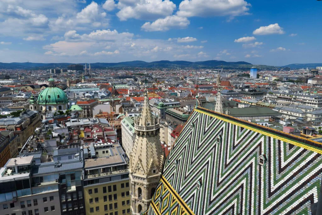 Luftaufnahme von Wien mit dem bunten, gemusterten Dach des Stephansdoms im Vordergrund. Das Stadtbild umfasst historische und moderne Gebäude unter einem teilweise bewölkten Himmel mit fernen Hügeln am Horizont.